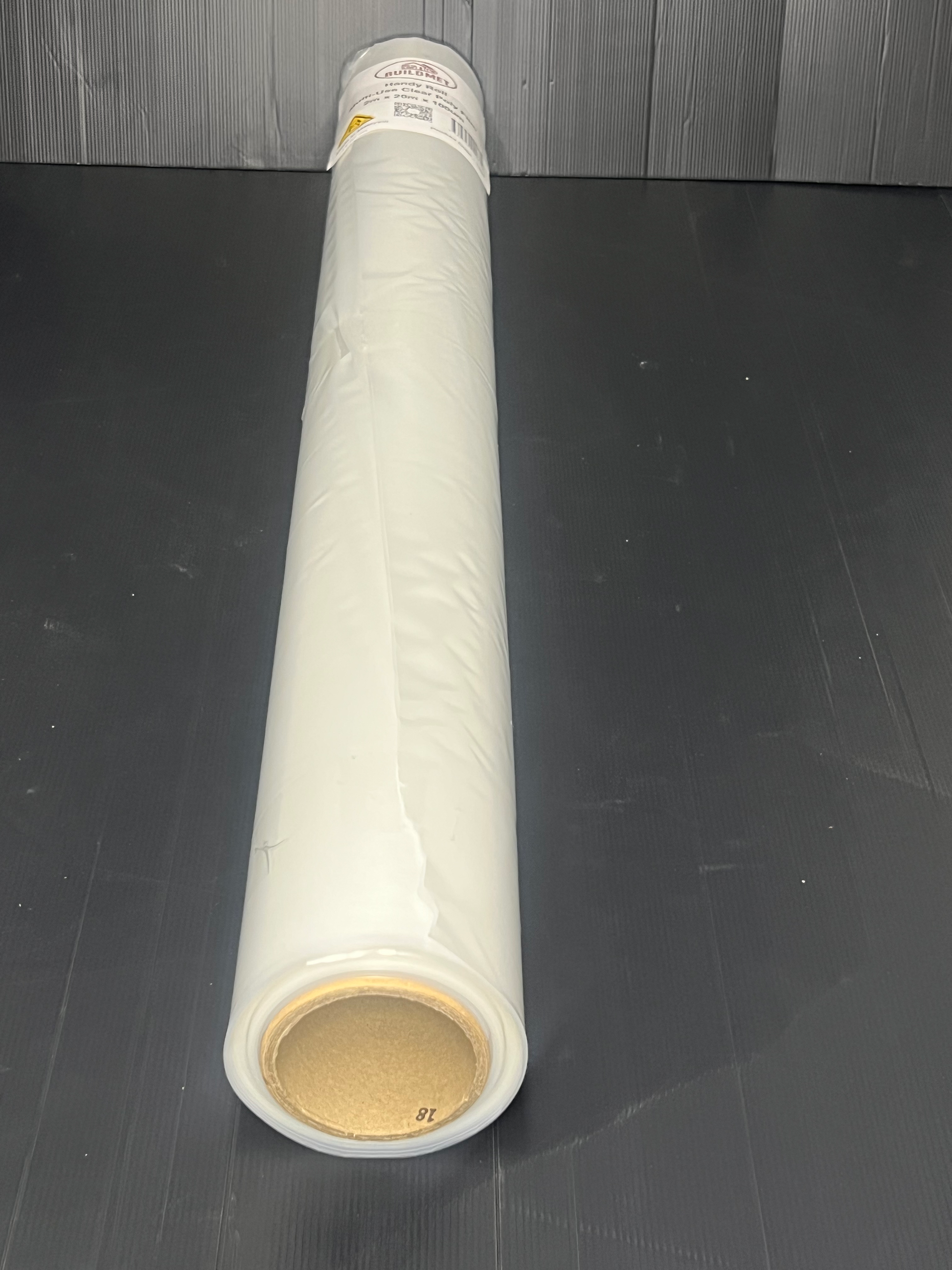 A roll of Pacific Packaging Australia's Buildmet Clear Builders film laying flat on a dark grey floor with grey wood wall in the background.